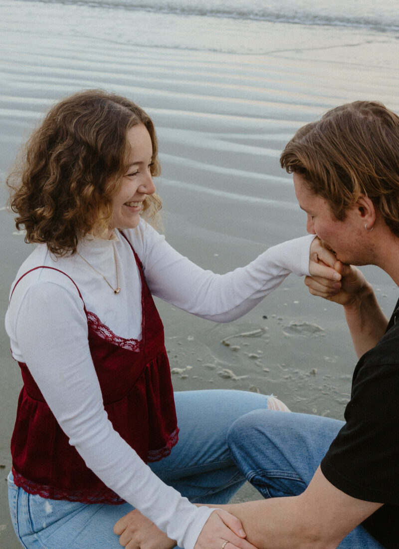 Couple crouching on Chesterman Beach in Tofino, he's holding and kissing her hand.