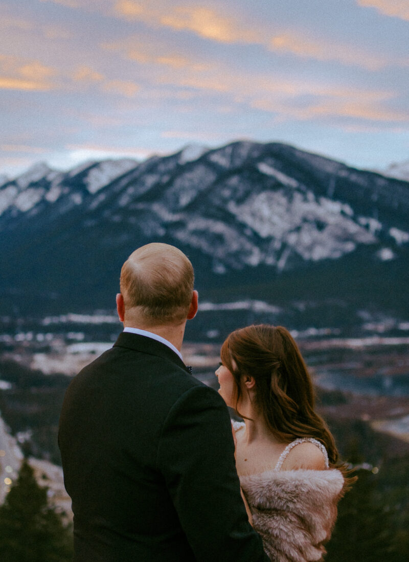 Bride and groom in embrace looking away at the mountain view at sunset on Mount Norquay.