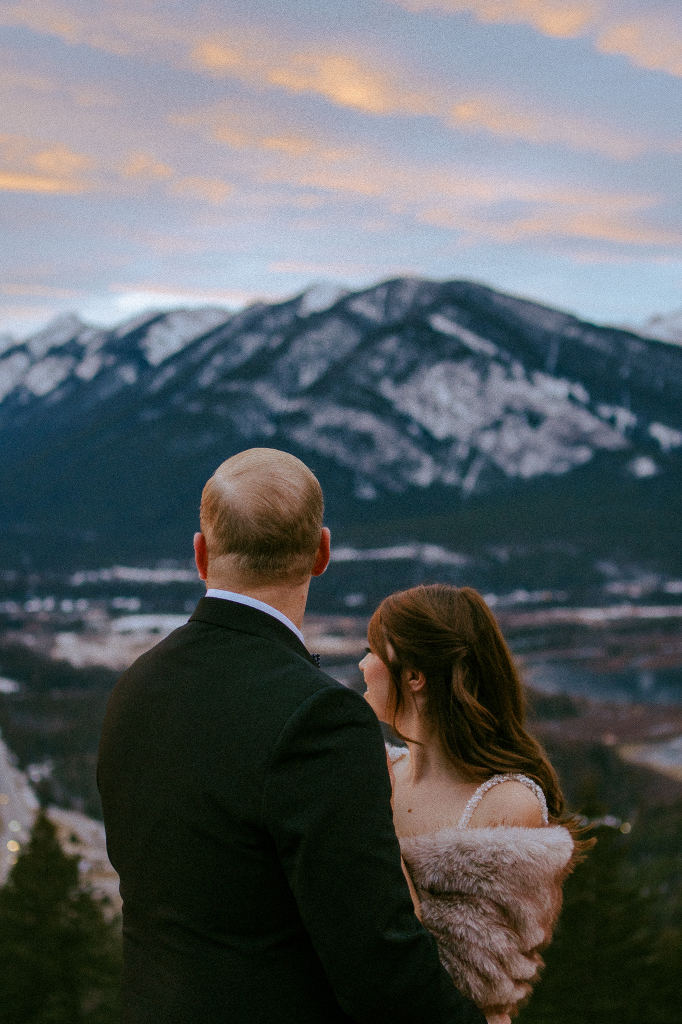Bride and groom in embrace looking away at the mountain view at sunset on Mount Norquay.