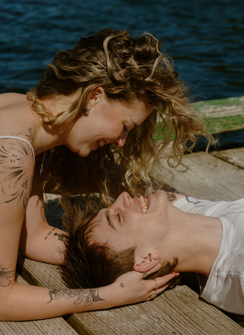 Couple shoot on a dock - He's lying on his back and she's propped up on her elbows looking down at him smiling.