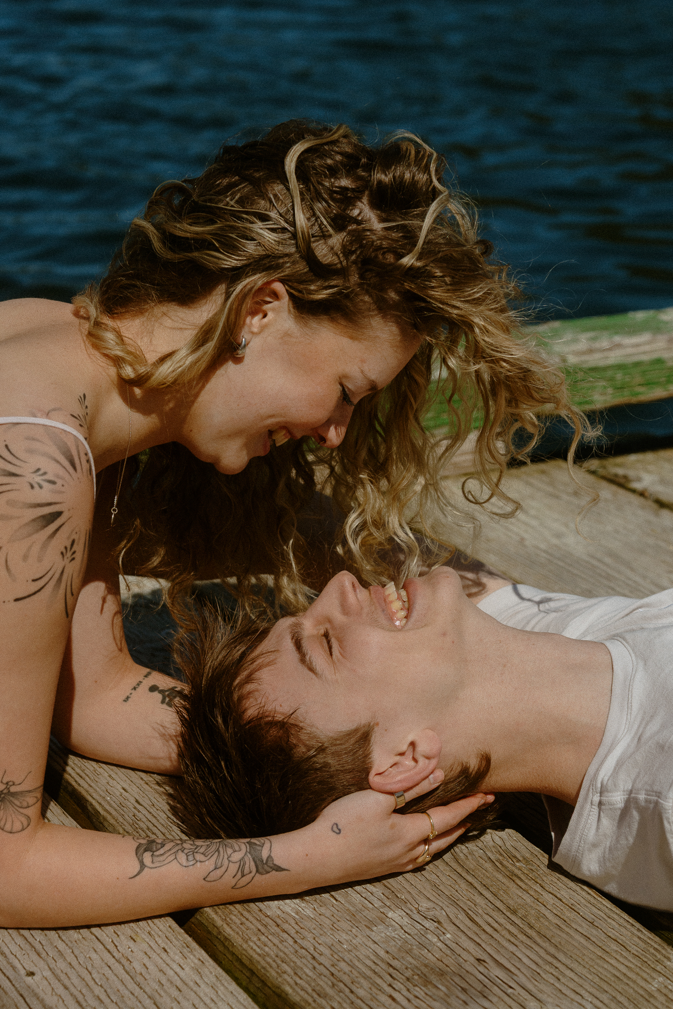 Couple shoot on a dock - He's lying on his back and she's propped up on her elbows looking down at him smiling.