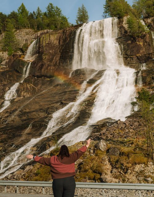 Fran running happily towards a waterfall with a rainbow, roadside in Norway